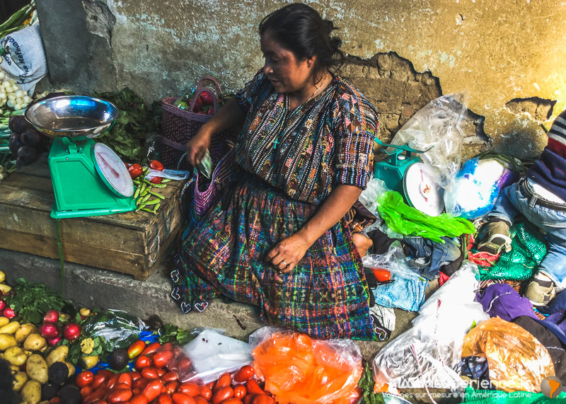 Femme en tenue traditionnelle vendant des légumes avec balance à un marché du Guatemala
