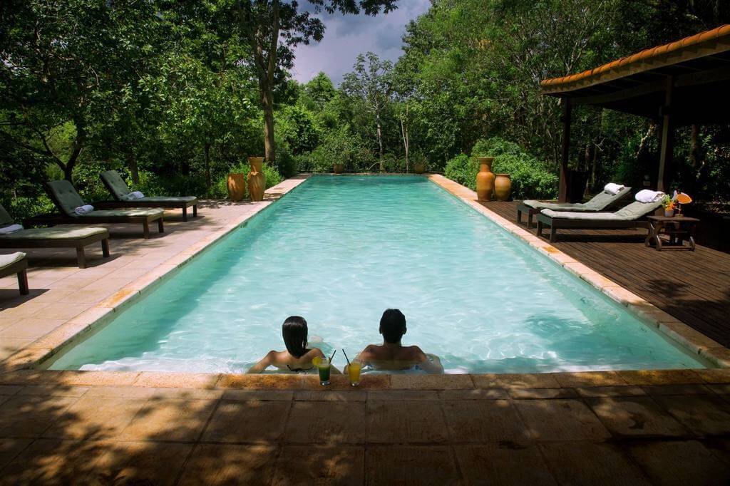 Piscine d'un lodge entourée d'arbres avec deux personnes se relaxant et cocktails