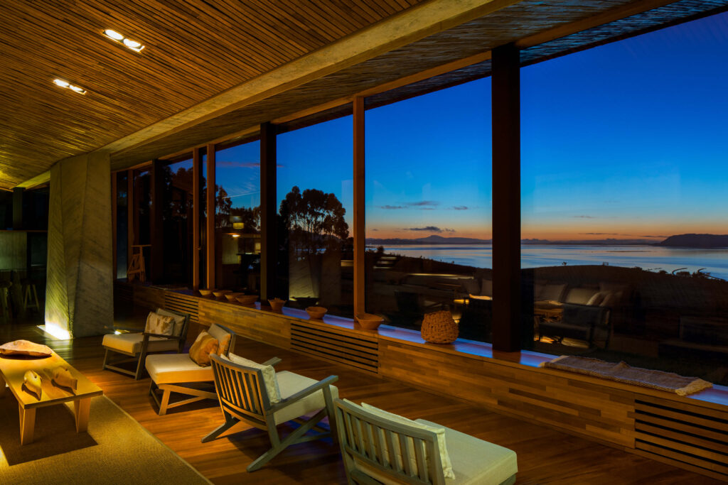 Salon d'hôtel en bois avec baies vitrées donnant sur la baie de Chiloé au crépuscule, Chili