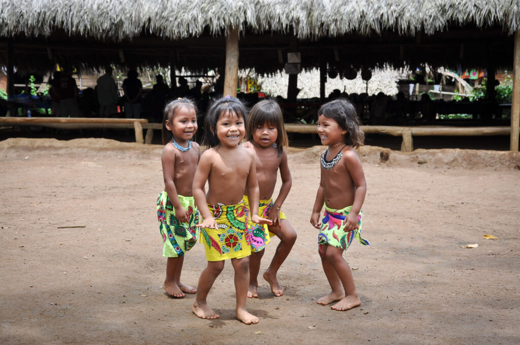 Quatre fillettes Embera en jupes colorées dansent et rient devant un village traditionnel au Panama