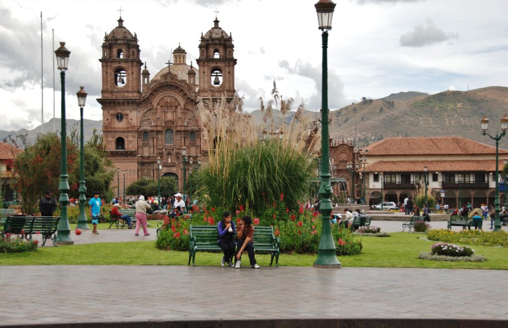 Place principale de Cusco avec église coloniale, jardins fleuris, bancs et passants, montagnes en arrière-plan