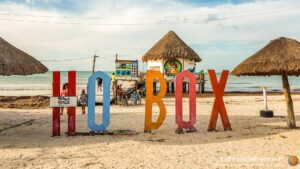 Lettres colorées HOLBOX sur plage de sable avec palapas et océan