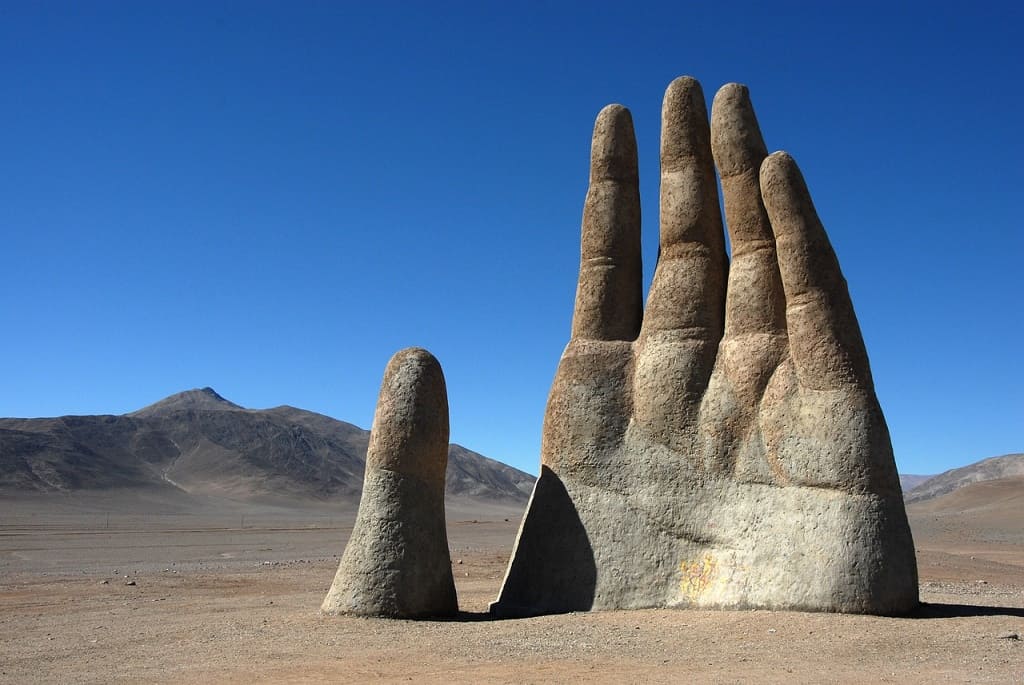 Sculpture géante d'une main émergeant du désert d'Atacama, montagnes en arrière-plan