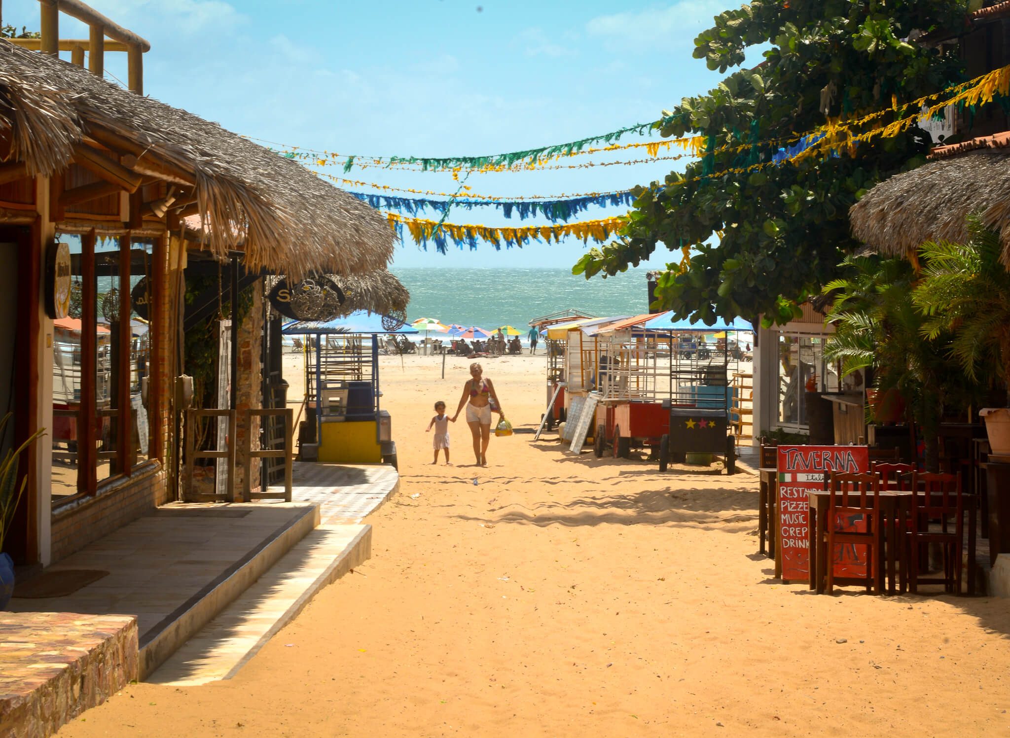 Rue de sable avec restaurants et cabanes de plage à Jericoacoara, femme et enfant marchant vers l'océan
