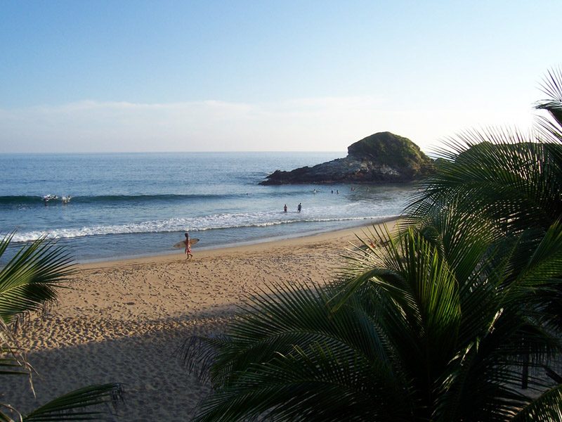 Plage mexicaine du Pacifique en plein jour, surfeur, baigneurs et rocher, palmiers au premier plan