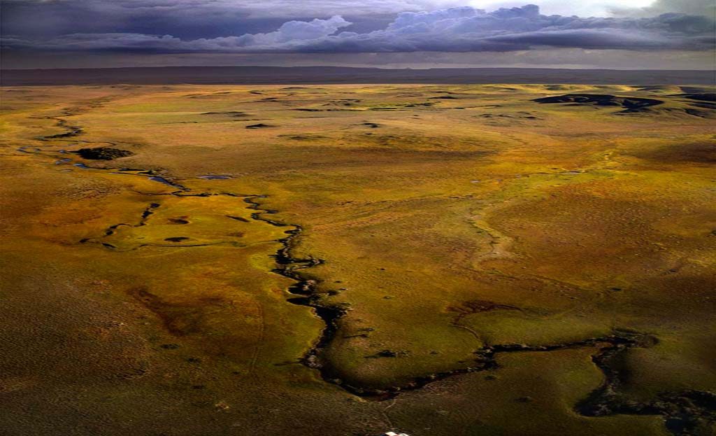 Vue aérienne d'une savane herbeuse traversée par un cours d'eau sinueux sous ciel orageux