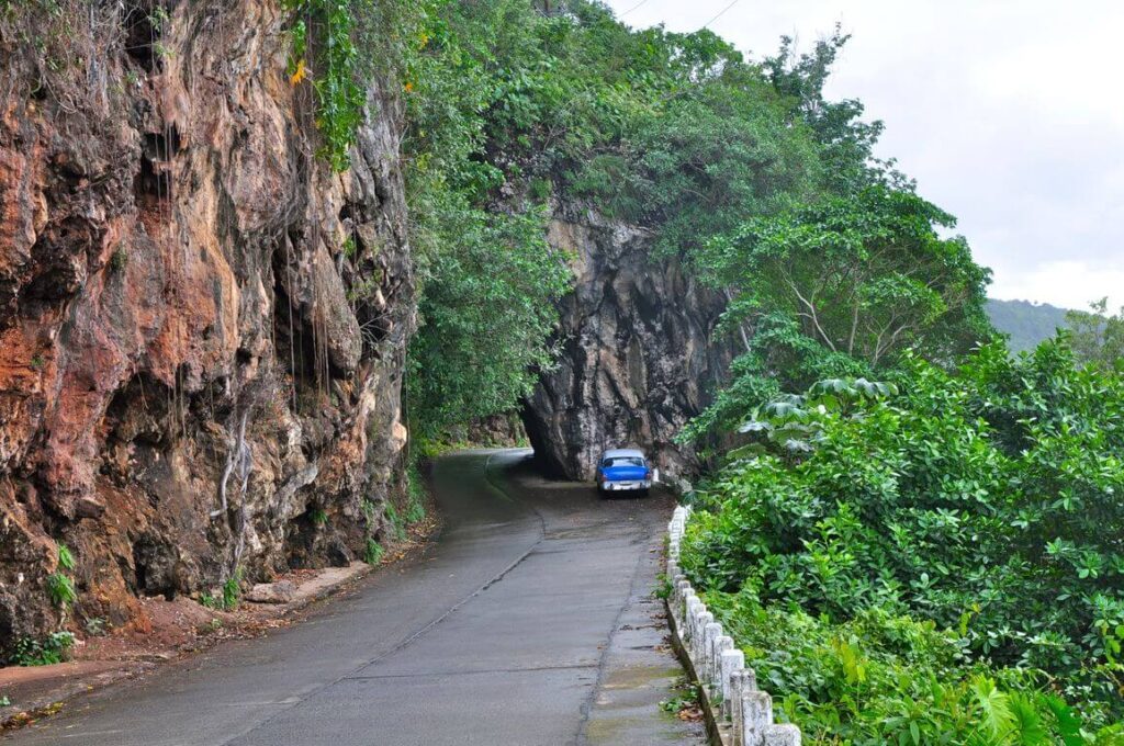 Voiture ancienne bleue sur route sinueuse entre falaise rocheuse et végétation, Baracoa, Cuba